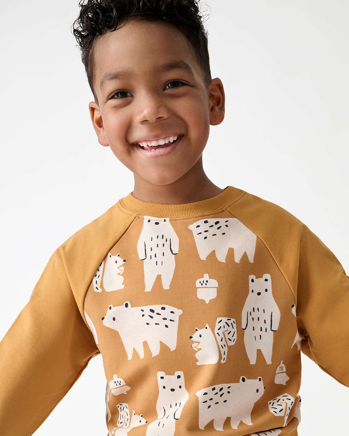 A smiling young boy wearing a mustard yellow long-sleeve sweatshirt with a playful white bear pattern, photographed against a clean white studio background.