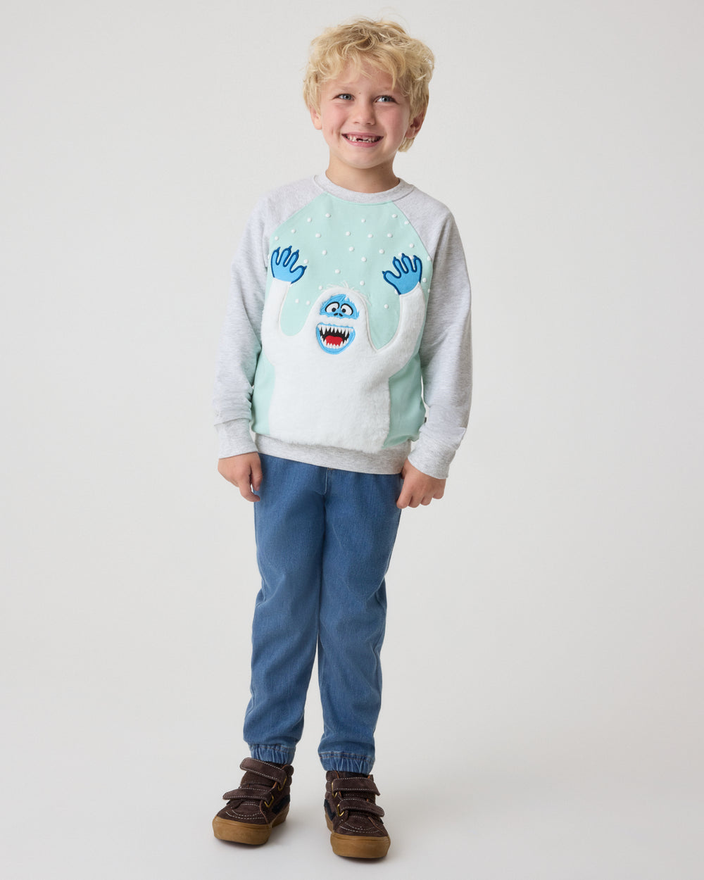 A young boy stands against a light gray studio background wearing a light gray and mint green sweatshirt with a blue cartoon character graphic, blue pants, and brown shoes.
