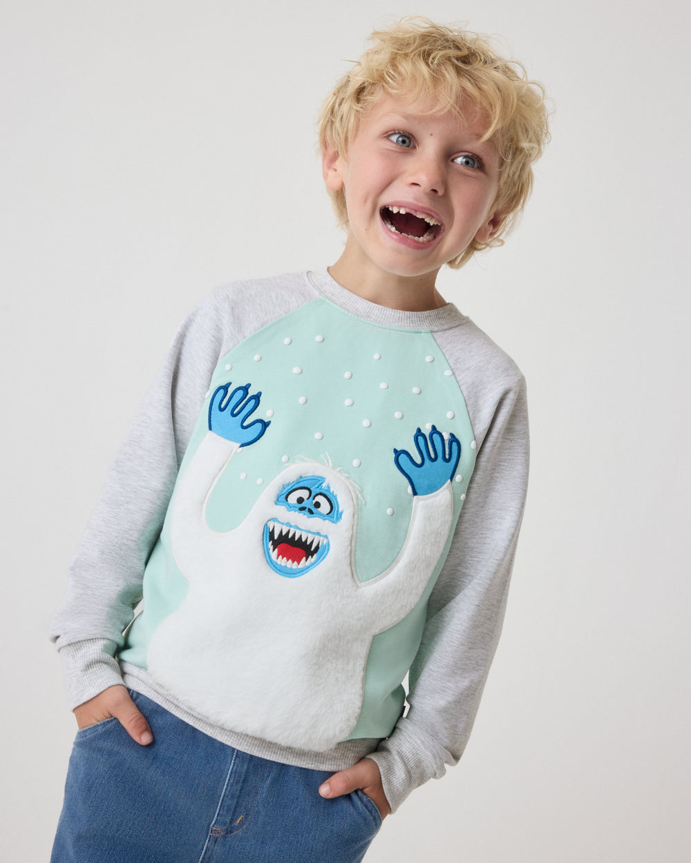 A young boy wearing a light gray and mint green long-sleeve sweatshirt with a blue monster graphic on the front, paired with blue jeans, against a plain light gray background.