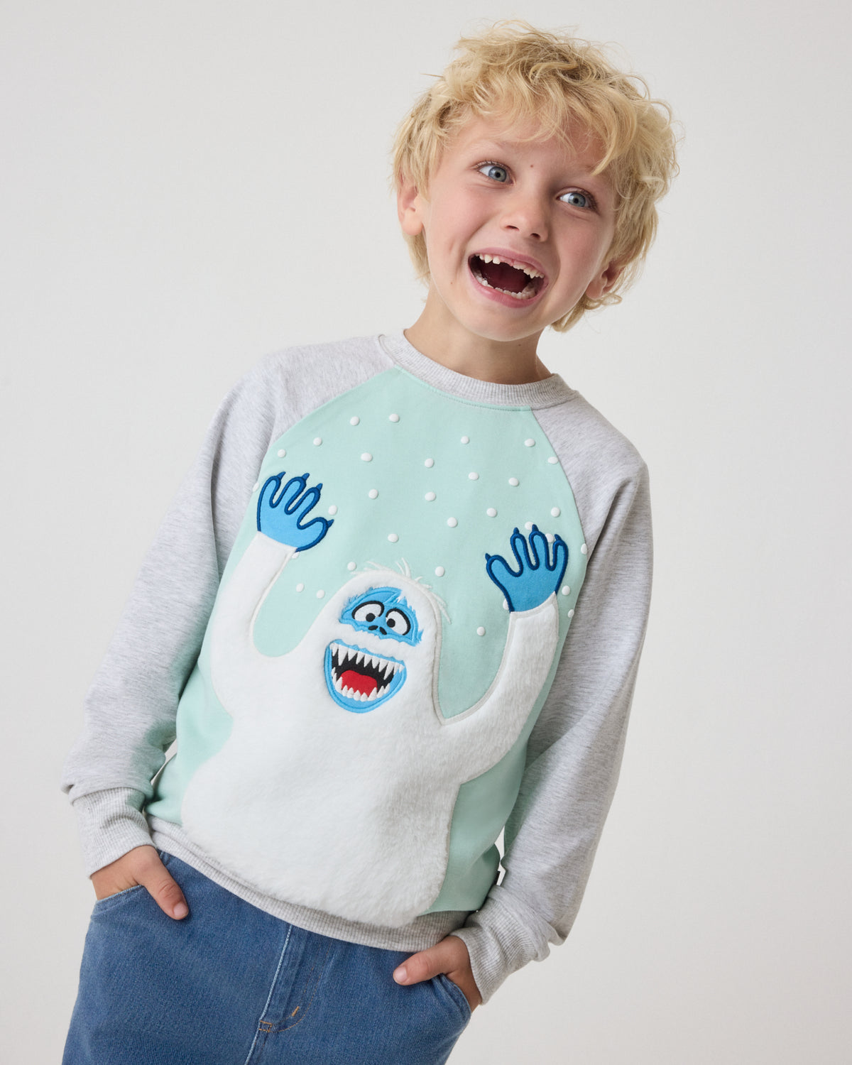 A young boy wearing a light gray and mint green long-sleeve sweatshirt with a blue monster graphic on the front, paired with blue jeans, against a plain light gray background.