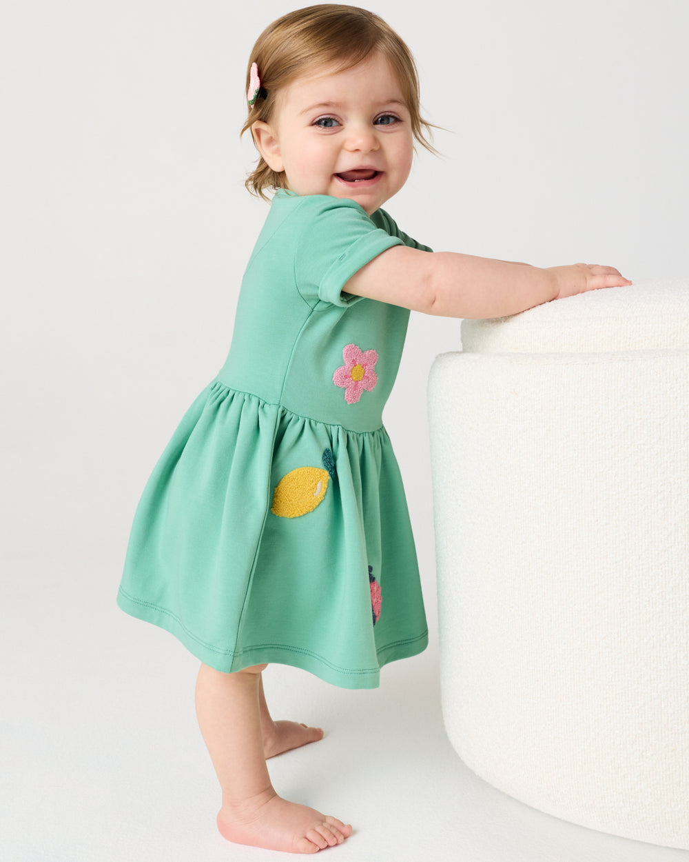 Baby wearing the Garden Party Dress with Bloomer holding onto a white ottoman