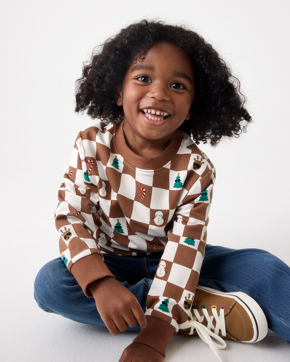 A smiling young child sits cross-legged on a light gray studio floor, wearing a brown and cream checkerboard long-sleeve sweatshirt with small holiday motifs, blue jeans, and tan sneakers.