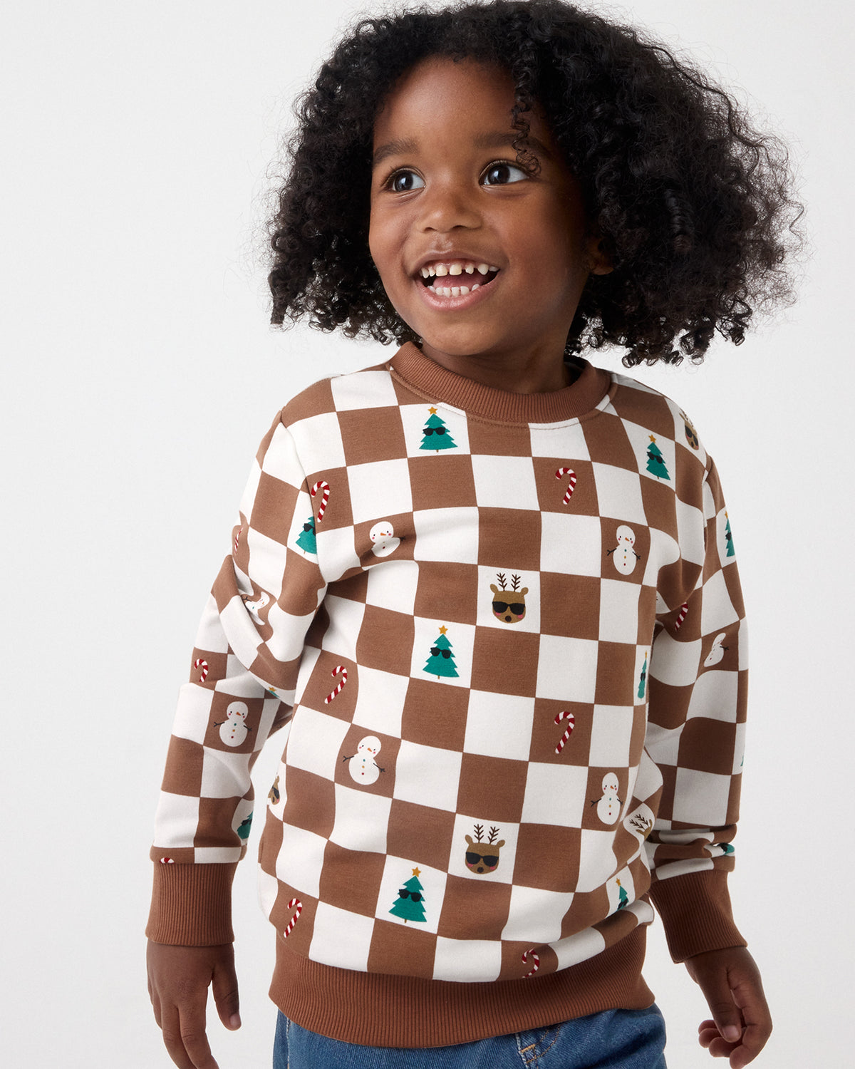 A smiling young child wearing a brown and cream checkerboard sweater decorated with Christmas motifs like trees, candy canes, snowmen, and gingerbread houses, paired with blue jeans against a plain light gray background.