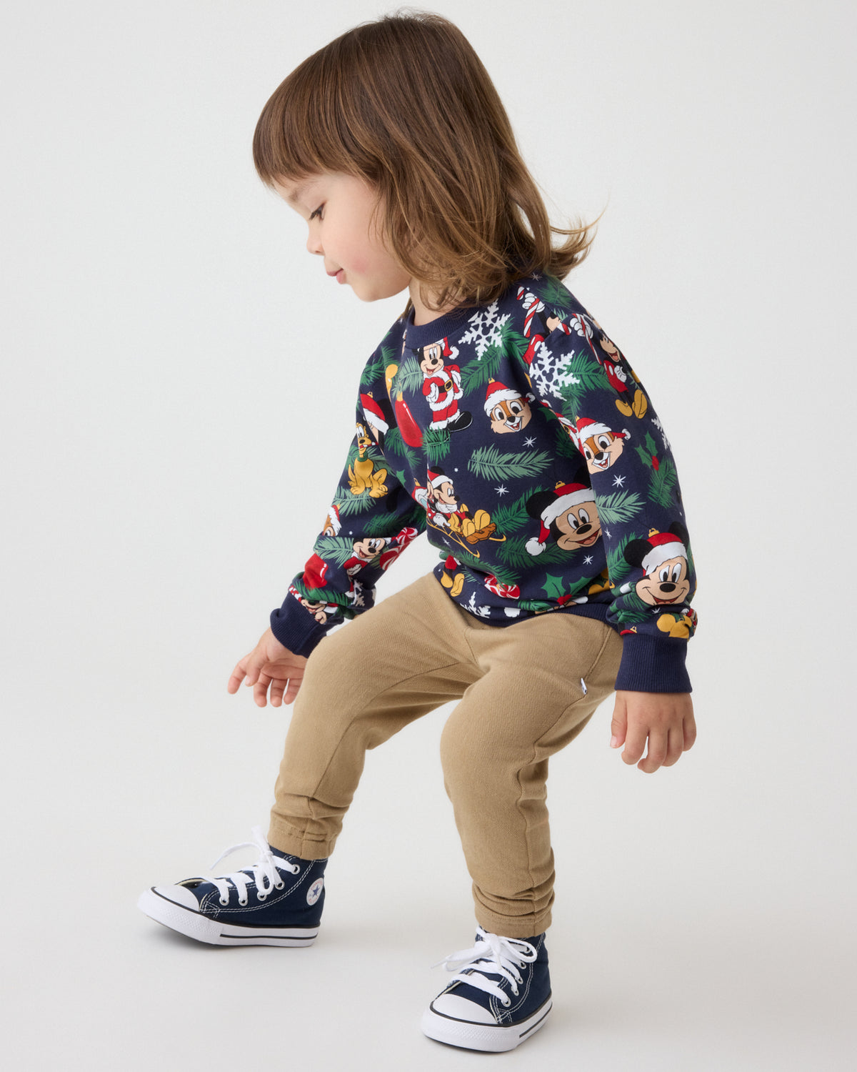 A toddler wearing a navy blue long-sleeve crewneck sweatshirt with a colorful Mickey Mouse Christmas print, paired with khaki pants and navy sneakers, captured against a clean white studio background.