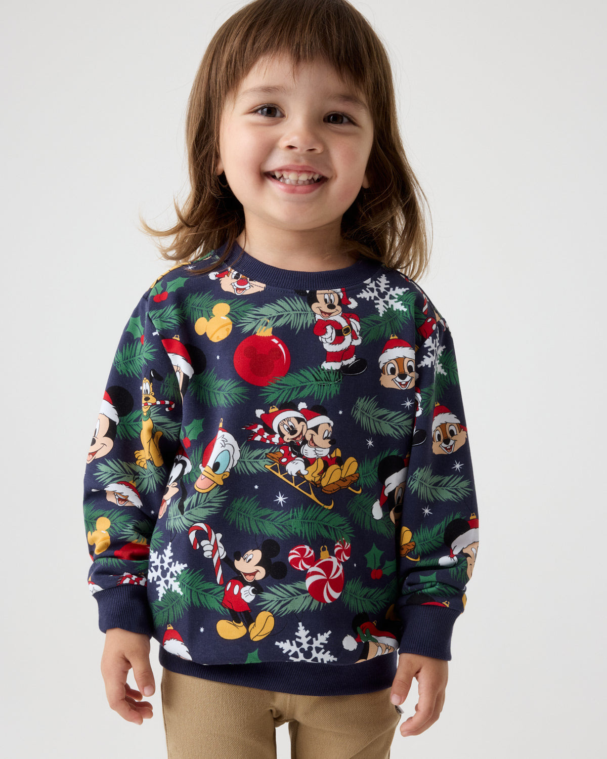 A young child wearing a navy blue Christmas crewneck sweatshirt with a festive cartoon print of Mickey Mouse and other Disney characters, paired with khaki pants, against a light gray studio background.