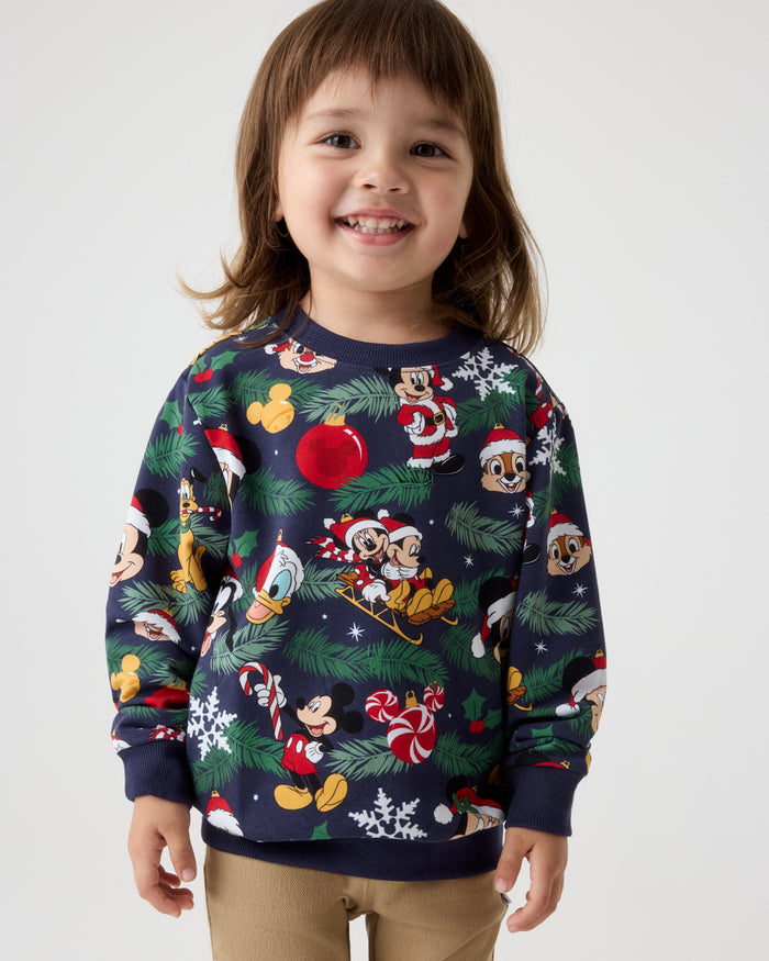 A young child wearing a navy blue Christmas crewneck sweatshirt with a festive cartoon print of Mickey Mouse and other Disney characters, paired with khaki pants, against a light gray studio background.