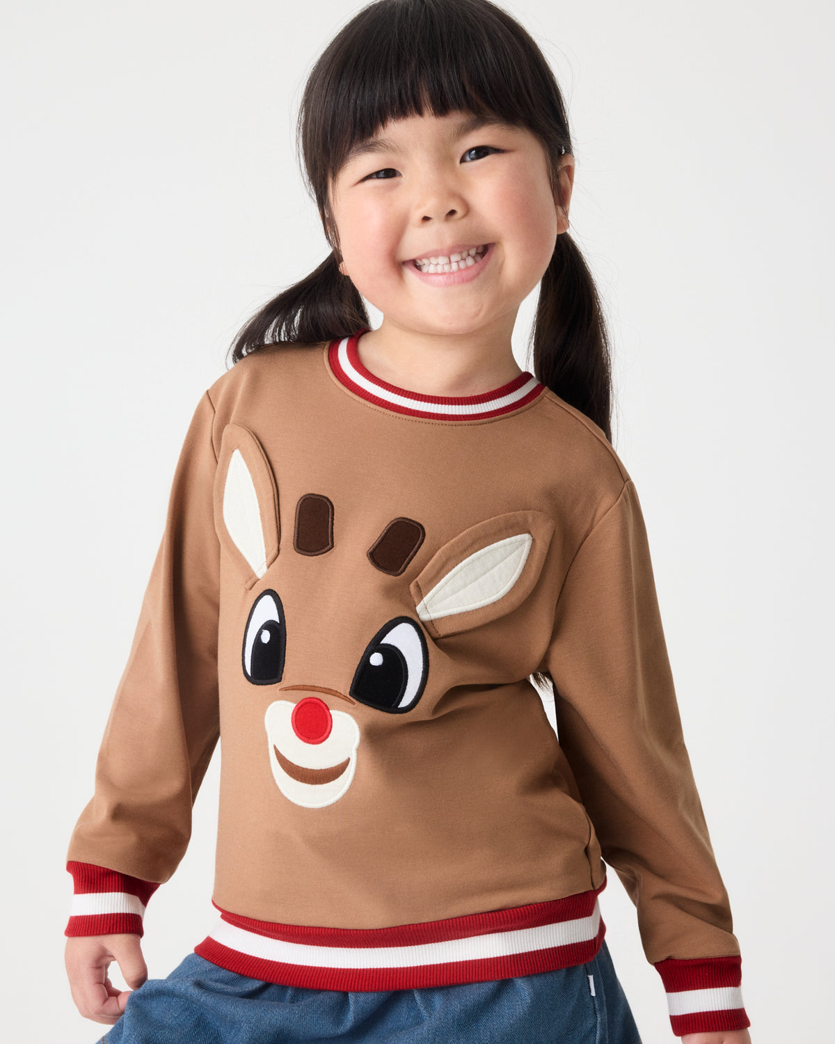 A young girl with dark hair in a ponytail smiling while wearing a brown long-sleeve sweatshirt with a reindeer face design and red and white striped accents, paired with blue jeans against a light gray background.
