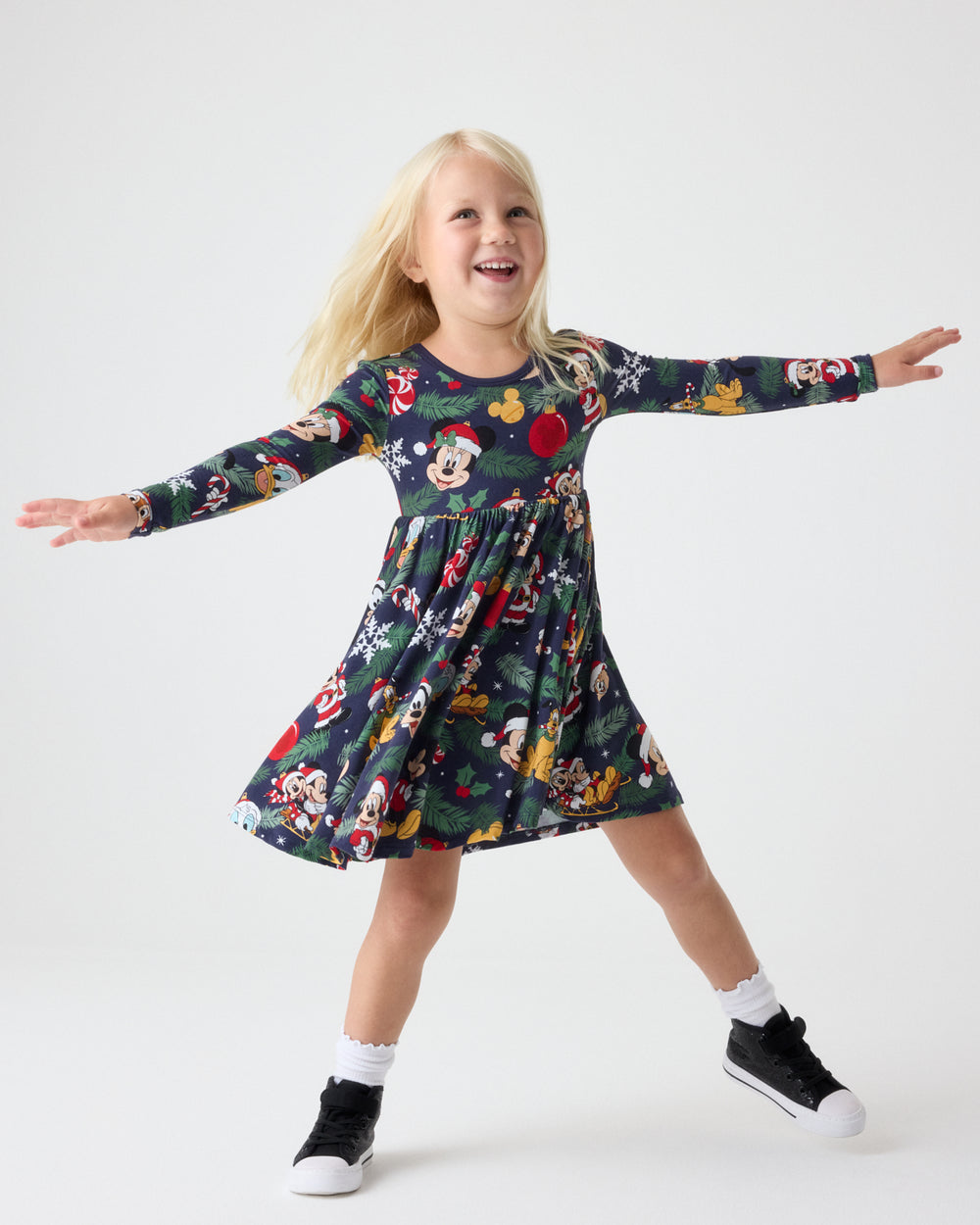 A young girl is smiling and posing with her arms outstretched, wearing a long-sleeve Mickey Mouse themed dress. The dress has a dark green base with a festive print of Mickey, Minnie, Donald, and Goofy in holiday attire, along with Christmas motifs like Santa and snowflakes.
