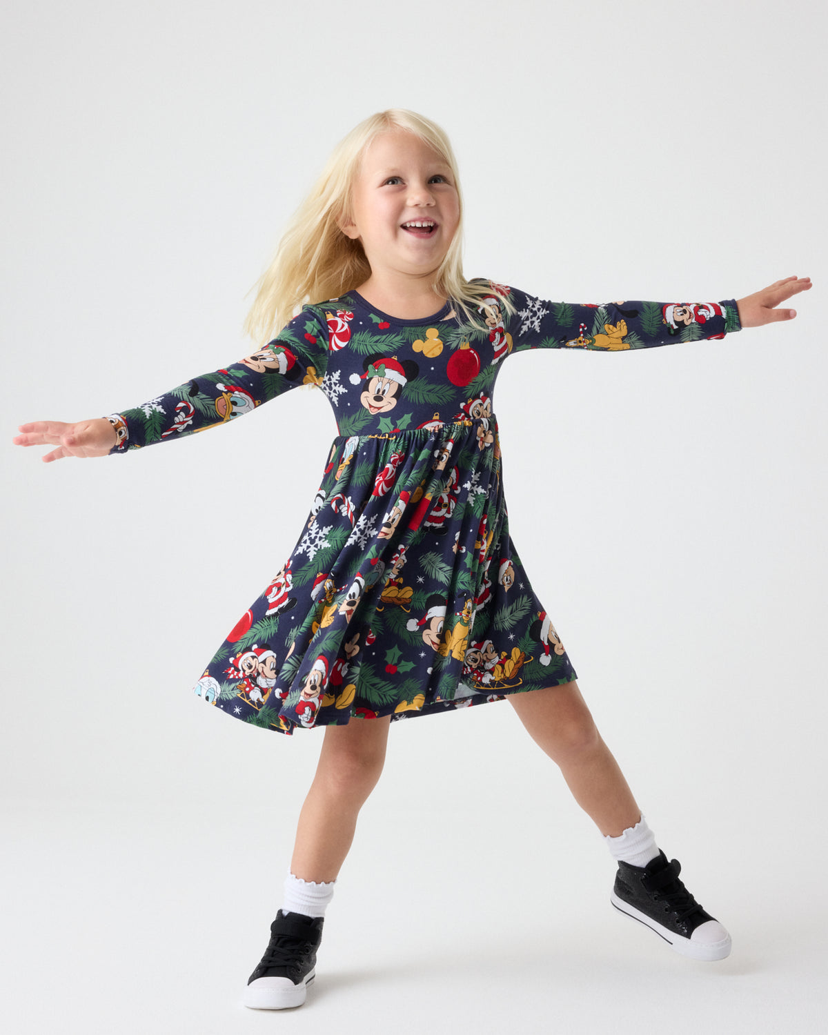 A young girl is smiling and posing with her arms outstretched, wearing a long-sleeve Mickey Mouse themed dress. The dress has a dark green base with a festive print of Mickey, Minnie, Donald, and Goofy in holiday attire, along with Christmas motifs like Santa and snowflakes.