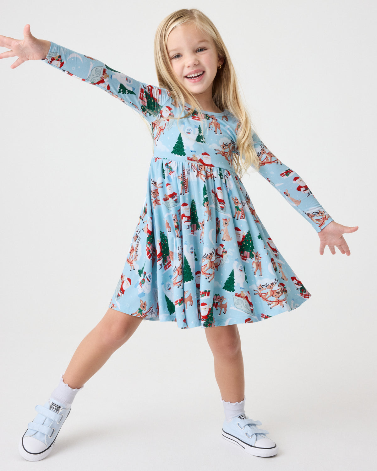 A young girl wearing a light blue long-sleeve dress with a festive Christmas print of Santa Claus, elves, and snowmen. She is posing with her arms outstretched on a seamless white studio background.