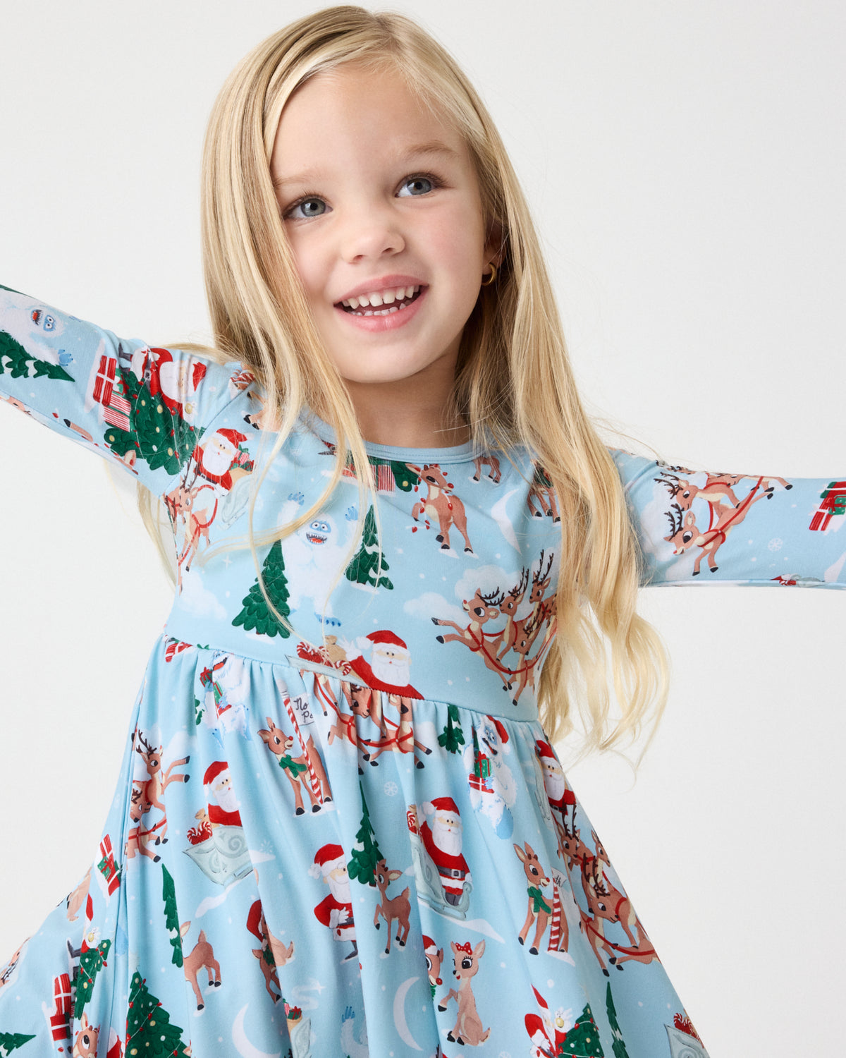 A young girl wearing a light blue long-sleeve dress with a festive Christmas print featuring Santa Claus, reindeer, and Christmas trees, set against a plain light gray background.