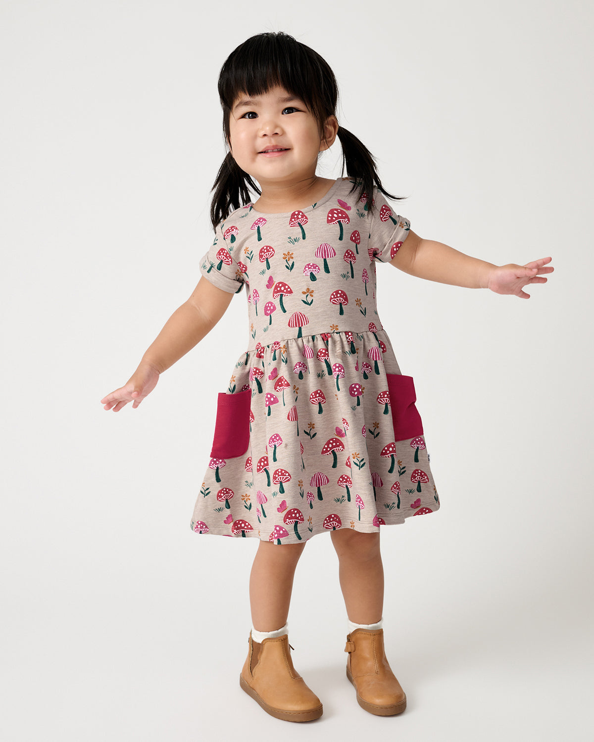 A young child wearing a short-sleeve dress with a pink mushroom print and magenta side pockets, standing against a light gray studio background.