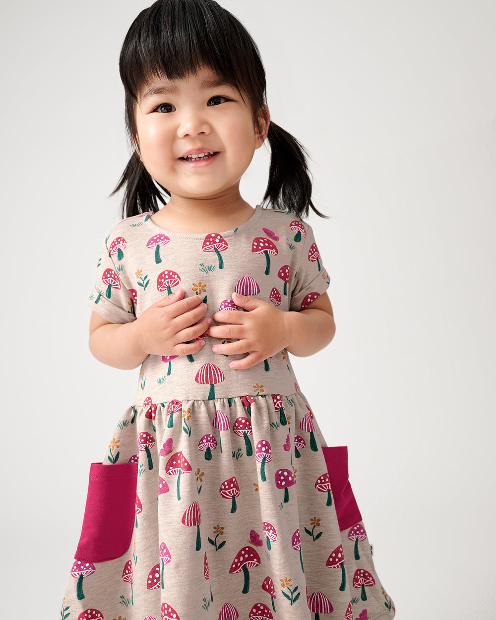 A young child wearing a short-sleeve dress with a pink and green mushroom print on a beige background, featuring solid pink side pockets, photographed against a light gray studio background.