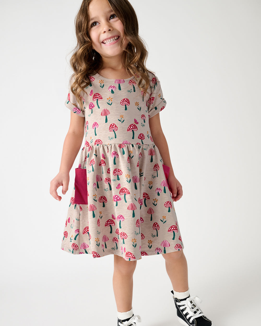 A young girl smiling and wearing a beige short-sleeve dress with a pink mushroom print and a solid pink pocket, paired with black and white sneakers, against a plain white studio background.