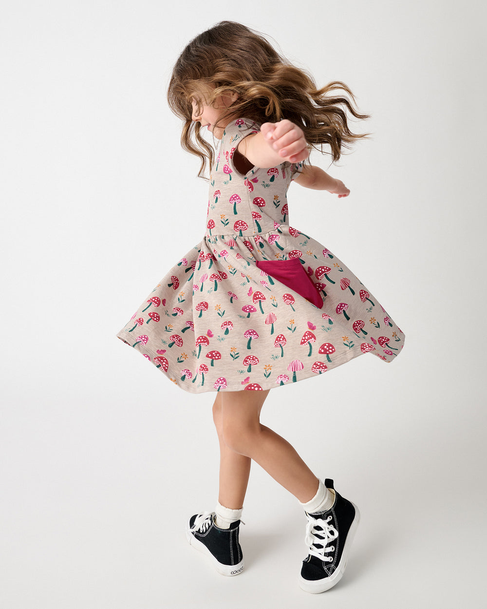 A young girl is captured mid-spin wearing a short-sleeve, knee-length dress with a pink mushroom print on a beige background. The dress features a large magenta pocket on the back side.