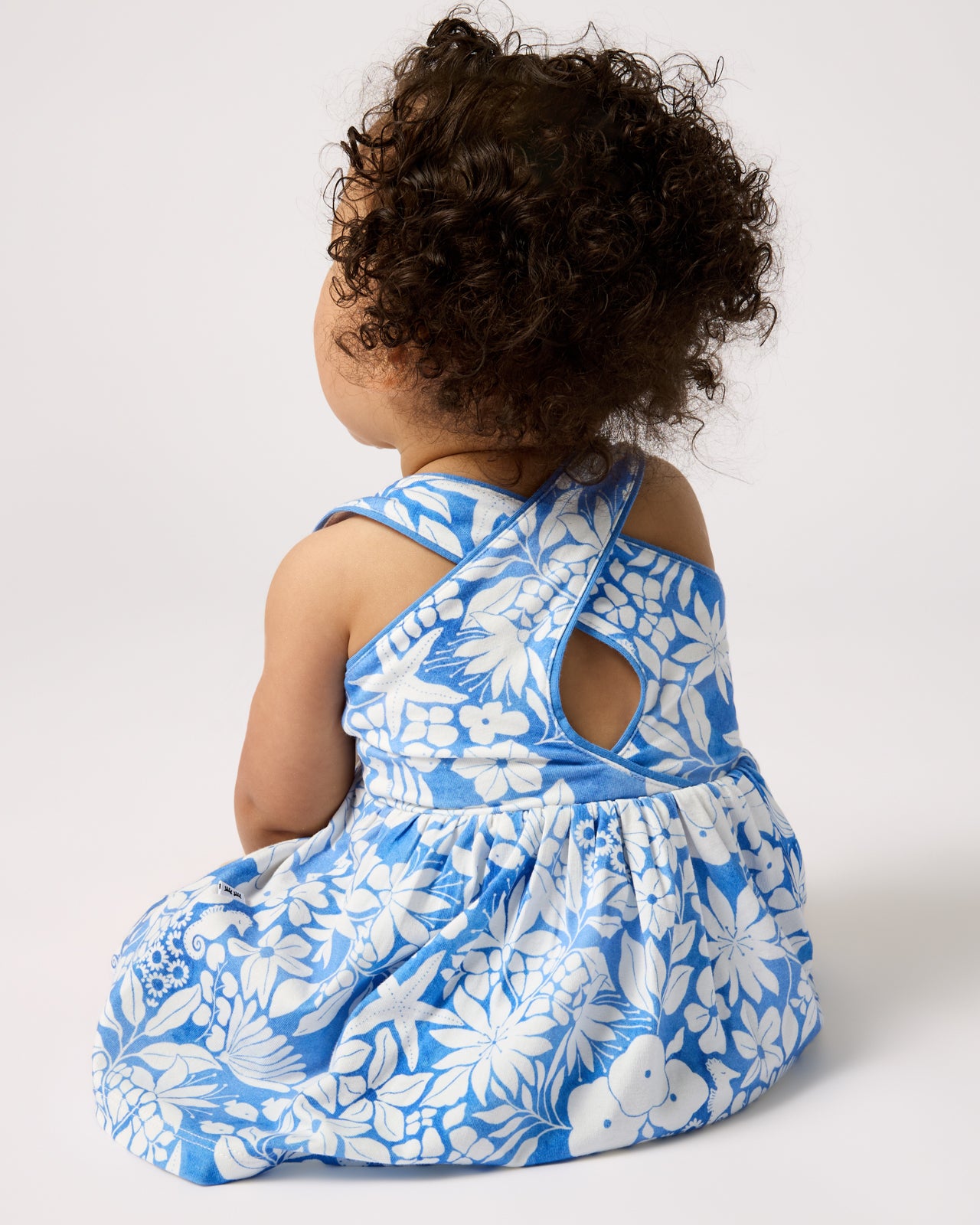 Back view of sitting baby wearing the Azure Garden Cross Back Skater Dress with Bloomer