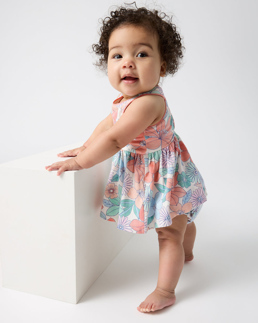 Baby holding onto a white block wearing the Shoreline Blooms Cross Back Skater Dress with Bloomer