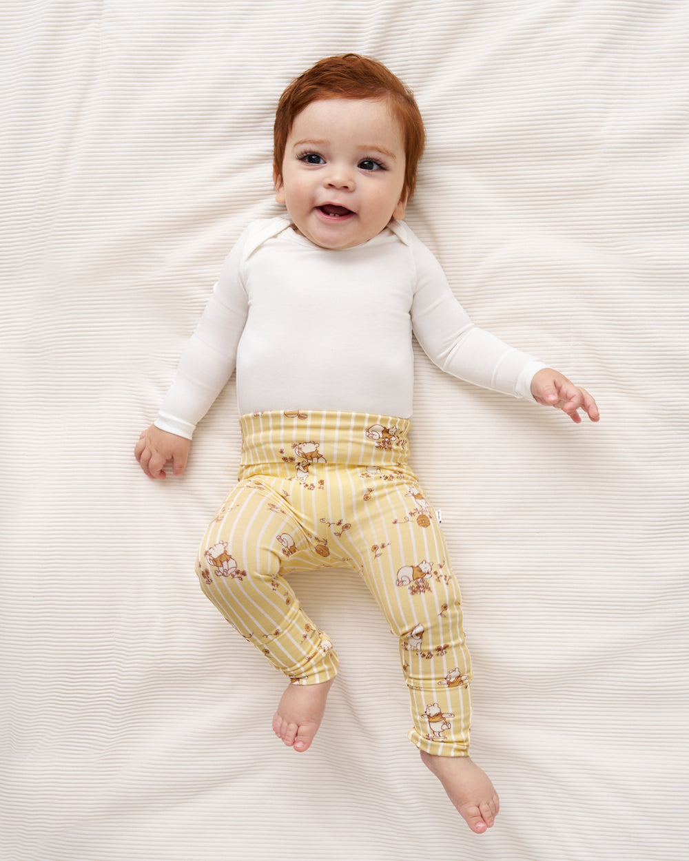 A baby lying on a cream-colored textured blanket, wearing a white long-sleeve top and yellow striped leggings with a small teddy bear pattern.
