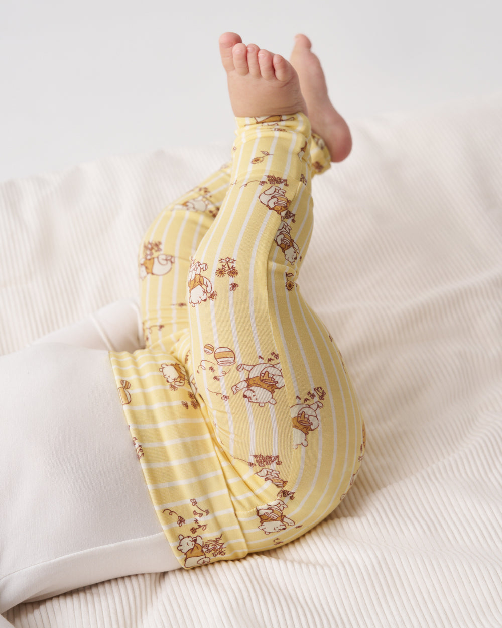 A baby's legs kicked up in the air while wearing yellow and white striped pajama pants with a brown cartoon bunny pattern, lying on a white ribbed blanket.