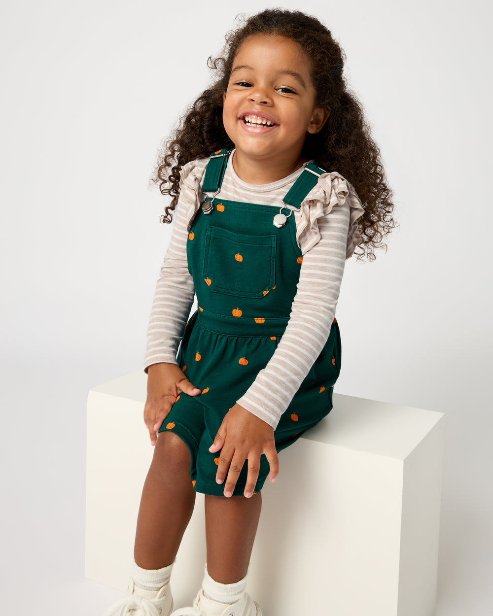 Sitting child wearing the Mini Pumpkins Skirt Overall Dress over a striped tee