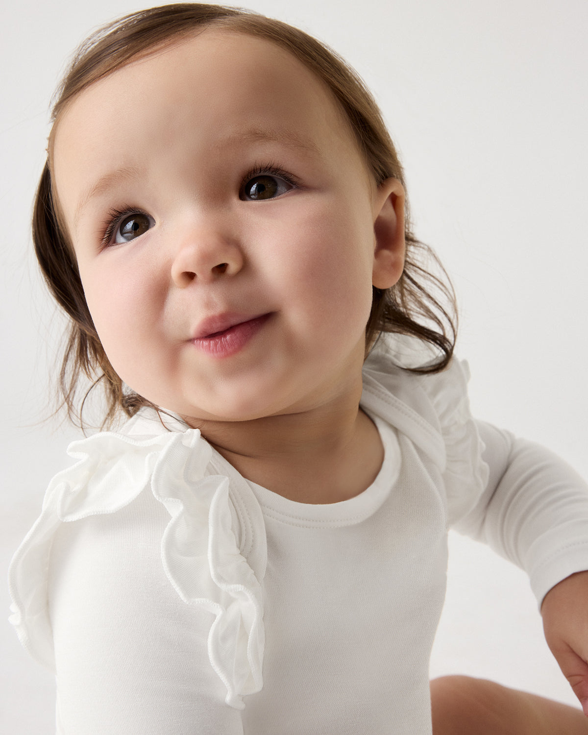 A close-up of a baby wearing a white long-sleeve bodysuit with a ruffled collar, photographed against a clean white background.