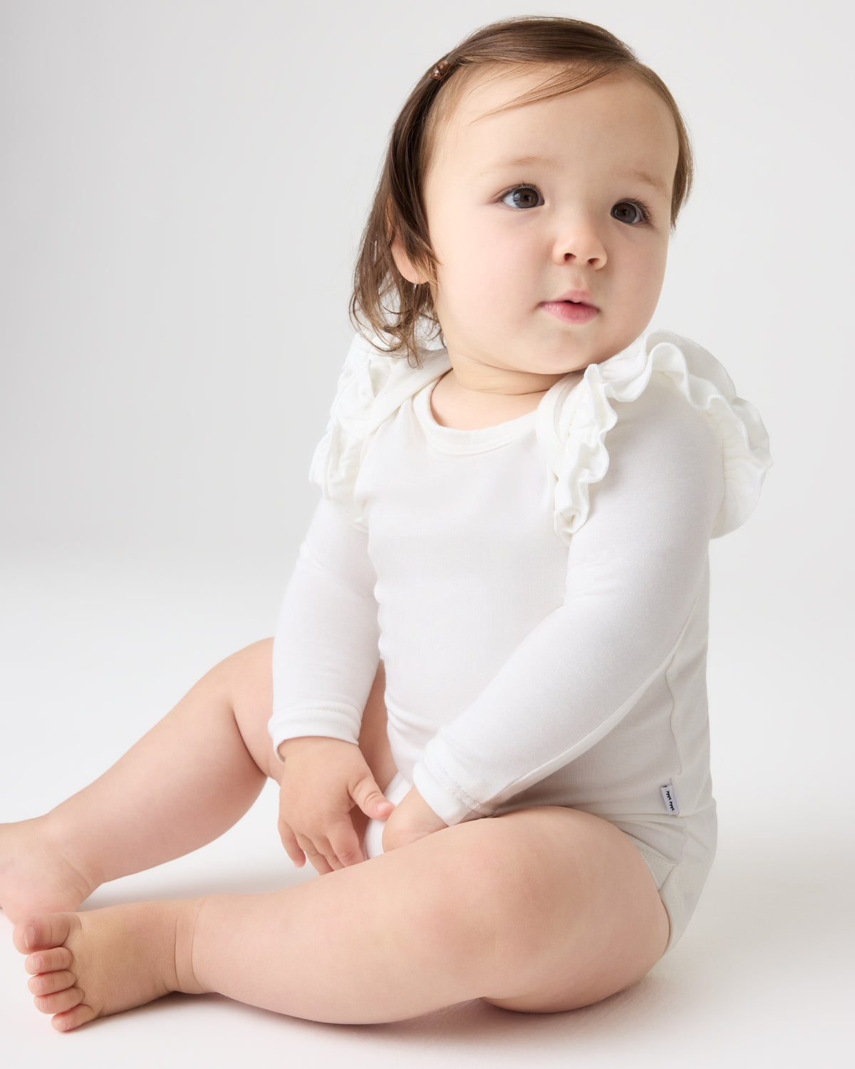 A baby sitting on a light gray studio background wearing a white long-sleeve bodysuit with ruffled shoulder details.