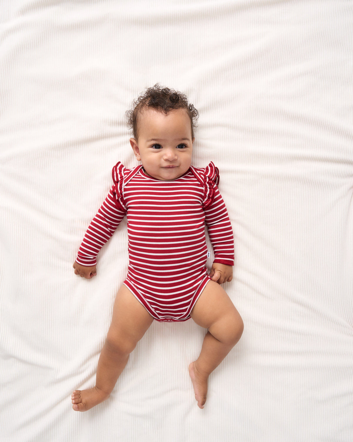 A baby lying on a white textured blanket, wearing a red and white horizontally striped long-sleeve bodysuit with ruffled shoulders.