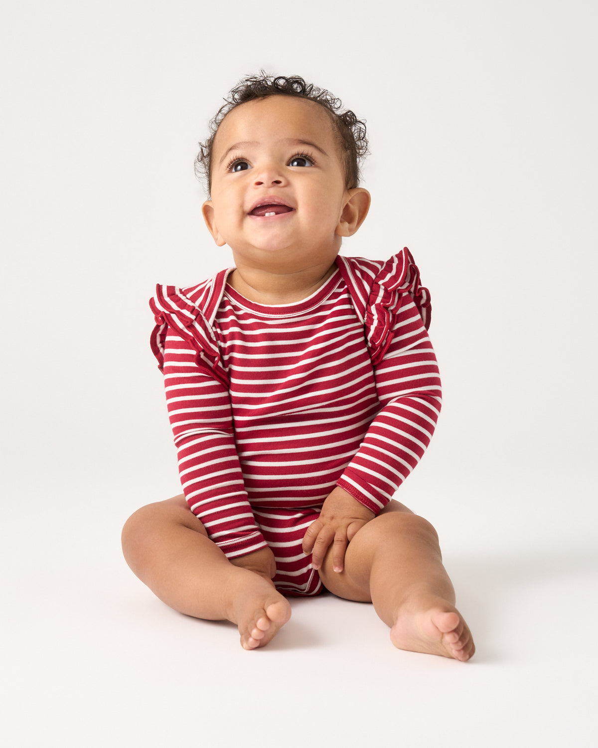 A smiling baby sitting on a white studio floor, wearing a red and white horizontally striped long-sleeve romper with ruffled shoulder details.