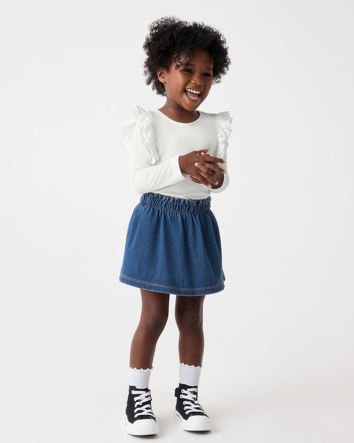 A young child stands against a light gray studio background, wearing a white long-sleeve top with ruffled shoulders, a blue denim mini skirt, white socks, and black-and-white sneakers.