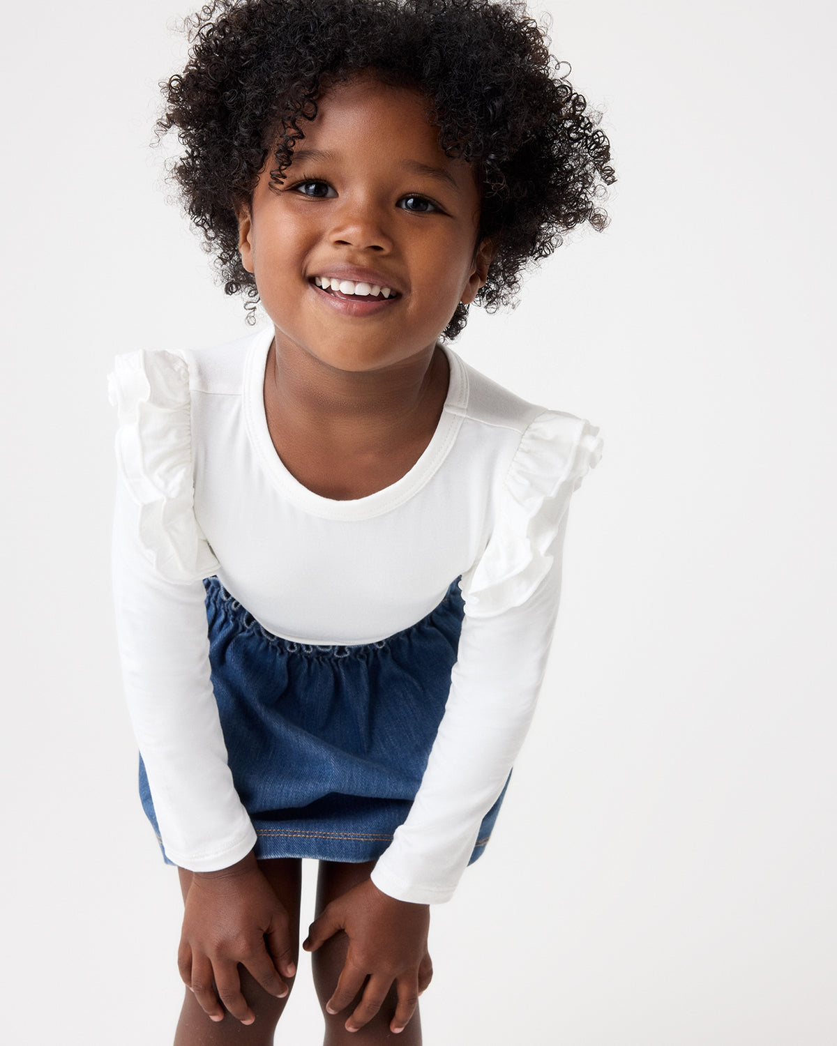 A young child smiling while wearing a white long-sleeve top with ruffled shoulders and a blue denim skirt, photographed against a plain white studio background.