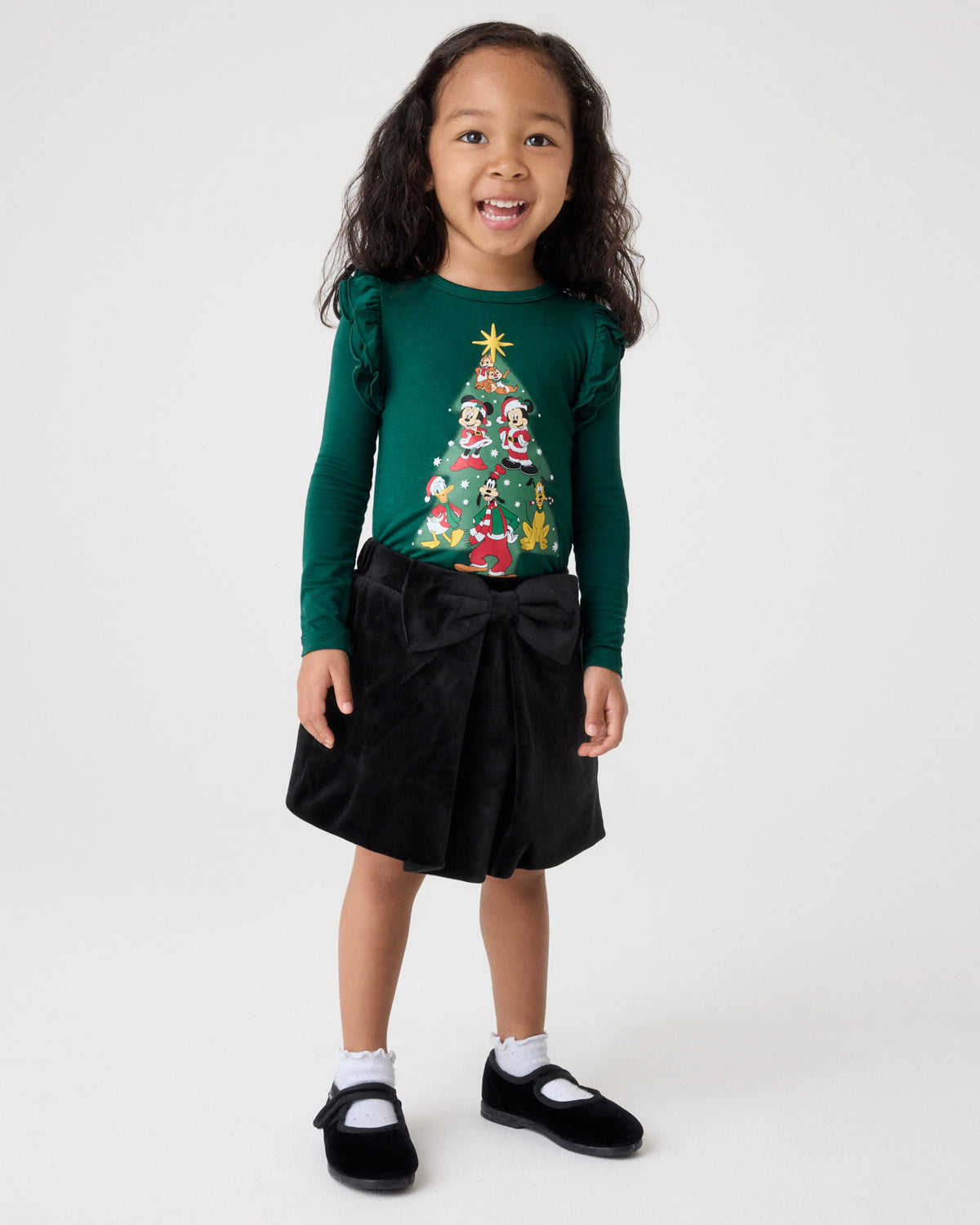 A young girl stands against a light gray studio background wearing a dark green long-sleeve top with a colorful Christmas tree graphic, a black skirt with a large bow, white socks, and black Mary Jane shoes.