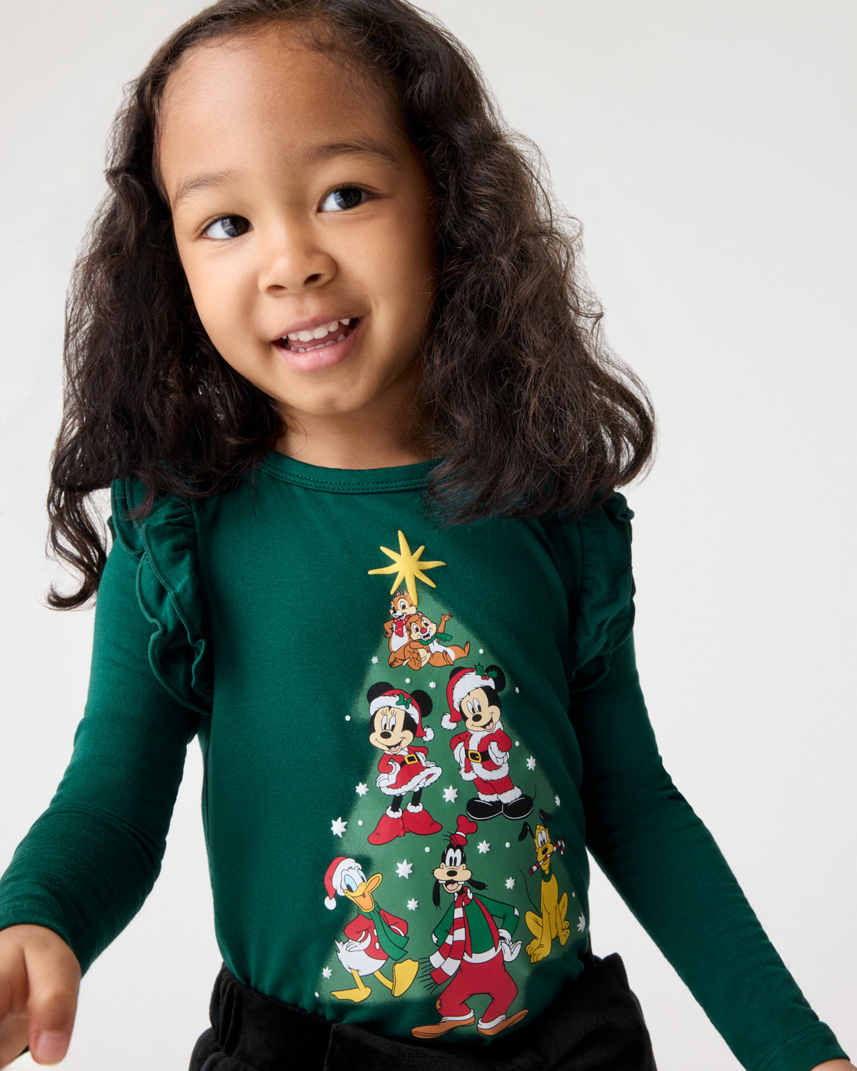 A young girl wearing a dark green long-sleeve shirt featuring a Christmas tree graphic with Disney characters, set against a plain light gray background.
