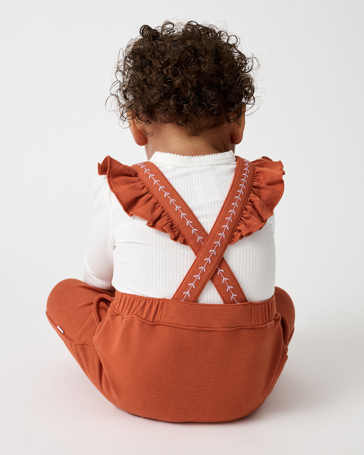 A baby seen from behind, wearing terracotta orange ruffle-shoulder baby overalls over a white ribbed long-sleeve top, sitting against a light gray background.