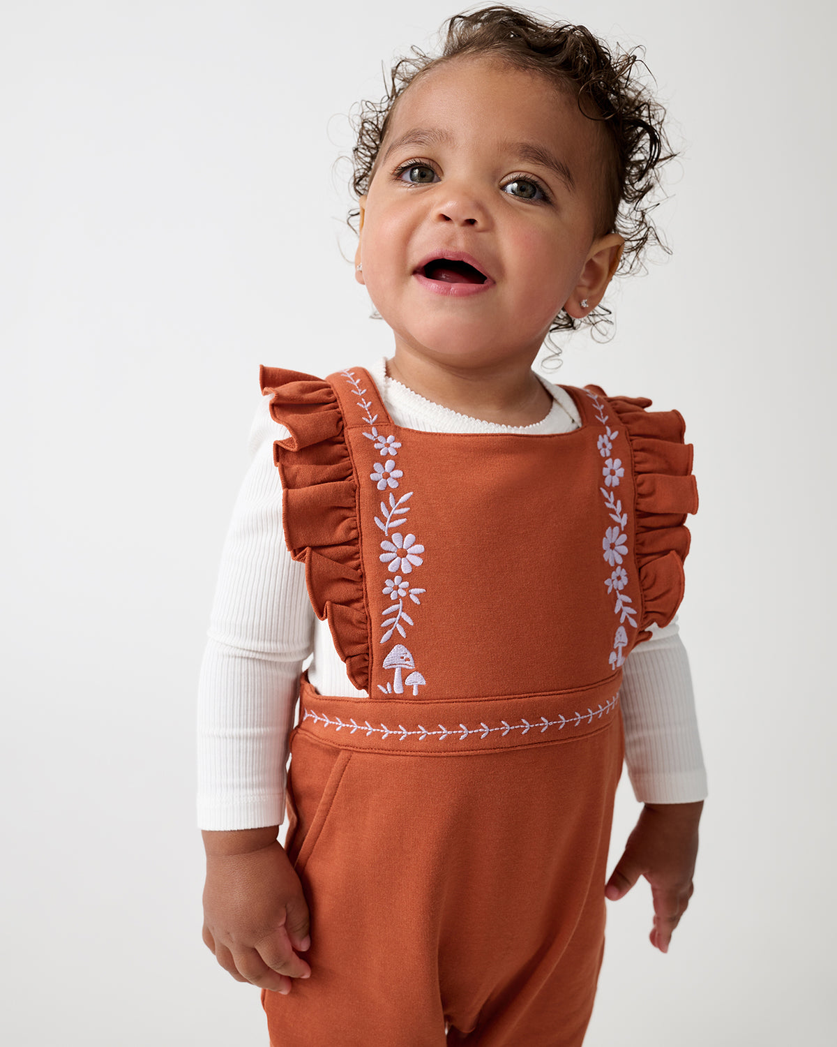 A toddler wearing a rust-orange overall romper with floral embroidery on the chest and sleeves, layered over a white long-sleeve top, photographed against a plain light gray background.