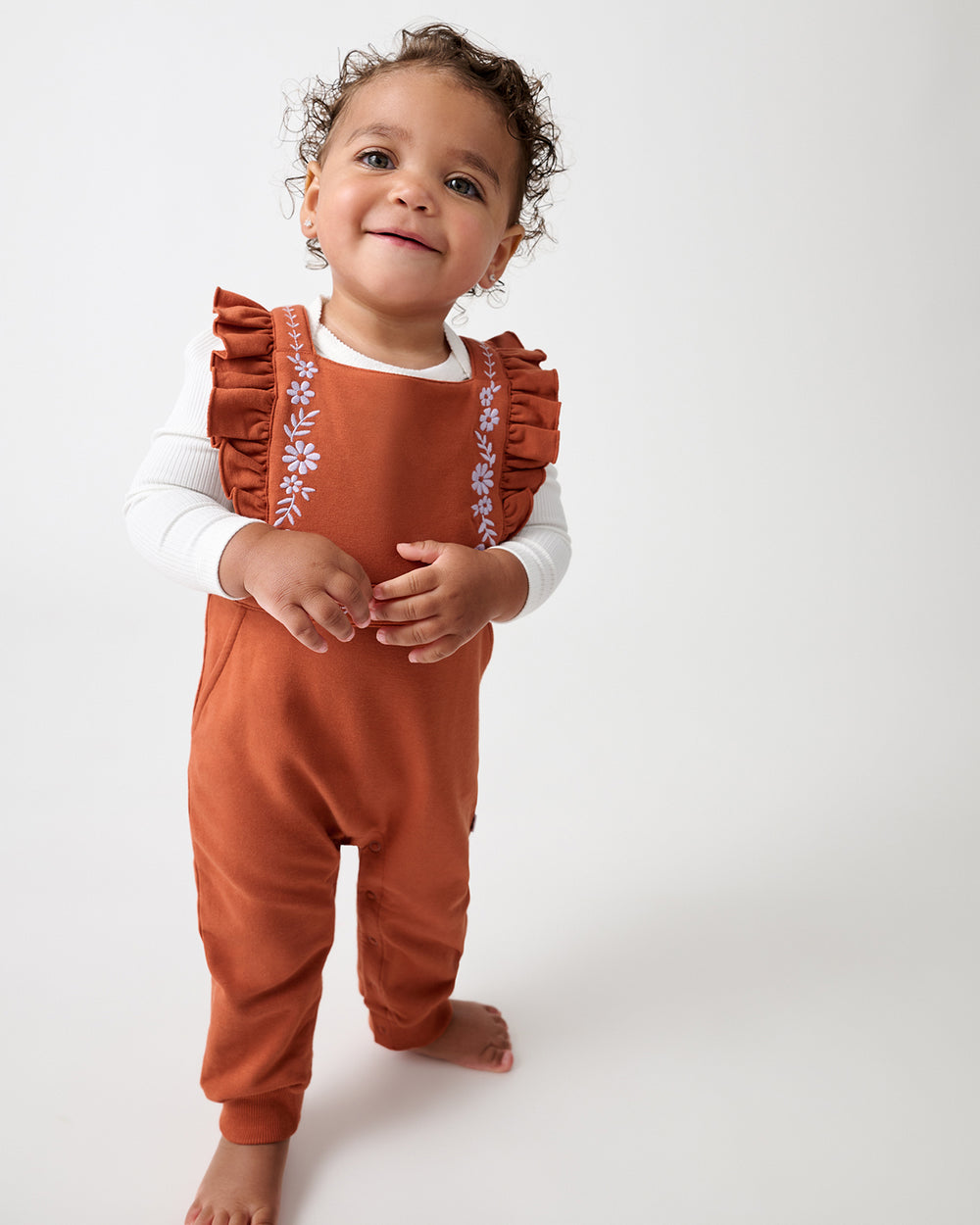 A toddler standing against a light gray studio background, wearing rust-orange overall-style baby clothes over a white long-sleeve top. The outfit has ruffled shoulder straps with small floral embroidery and a white ribbed collar.
