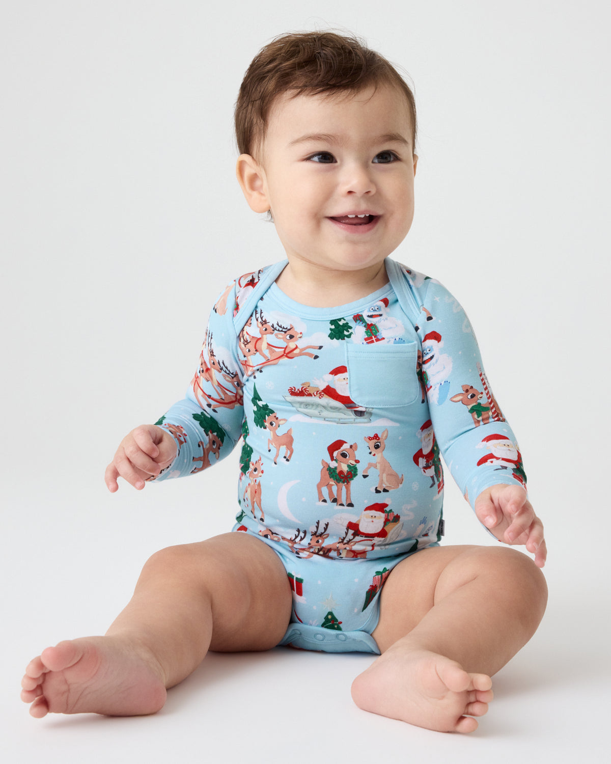 A smiling baby sitting on a white studio floor wearing a light blue long-sleeve romper with a festive Christmas Rudolph print.