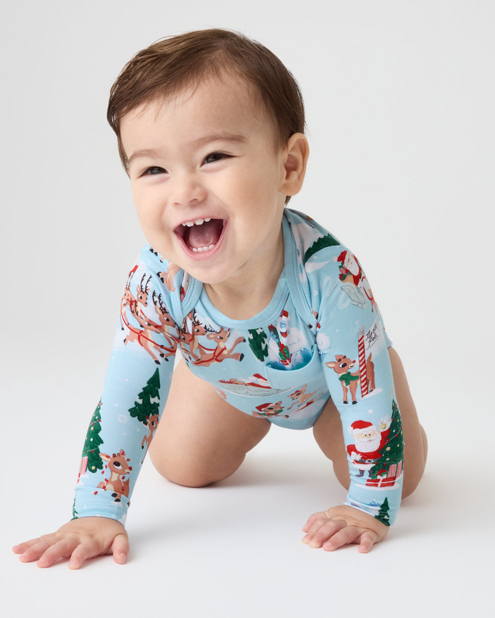 A smiling baby crawling on a white studio floor, wearing a light blue long-sleeve bodysuit with a Christmas print featuring Santa Claus, reindeer, trees, and snowflakes.