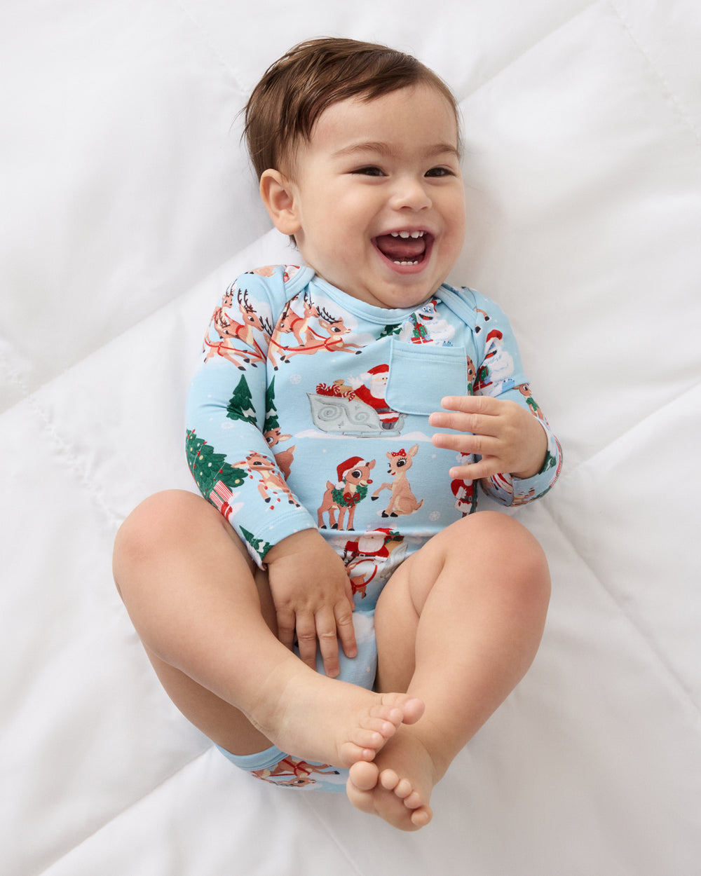 A smiling baby sitting on white bedding, wearing a light blue long-sleeve pajama top with a festive Christmas print featuring Santa, reindeer, and a tree.