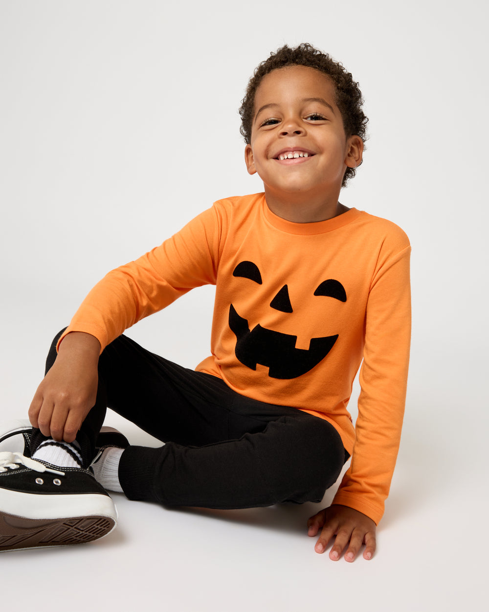 Sitting boy wearing the Jack-O'-Lantern Long Sleeve Relaxed Tee with black joggers 
