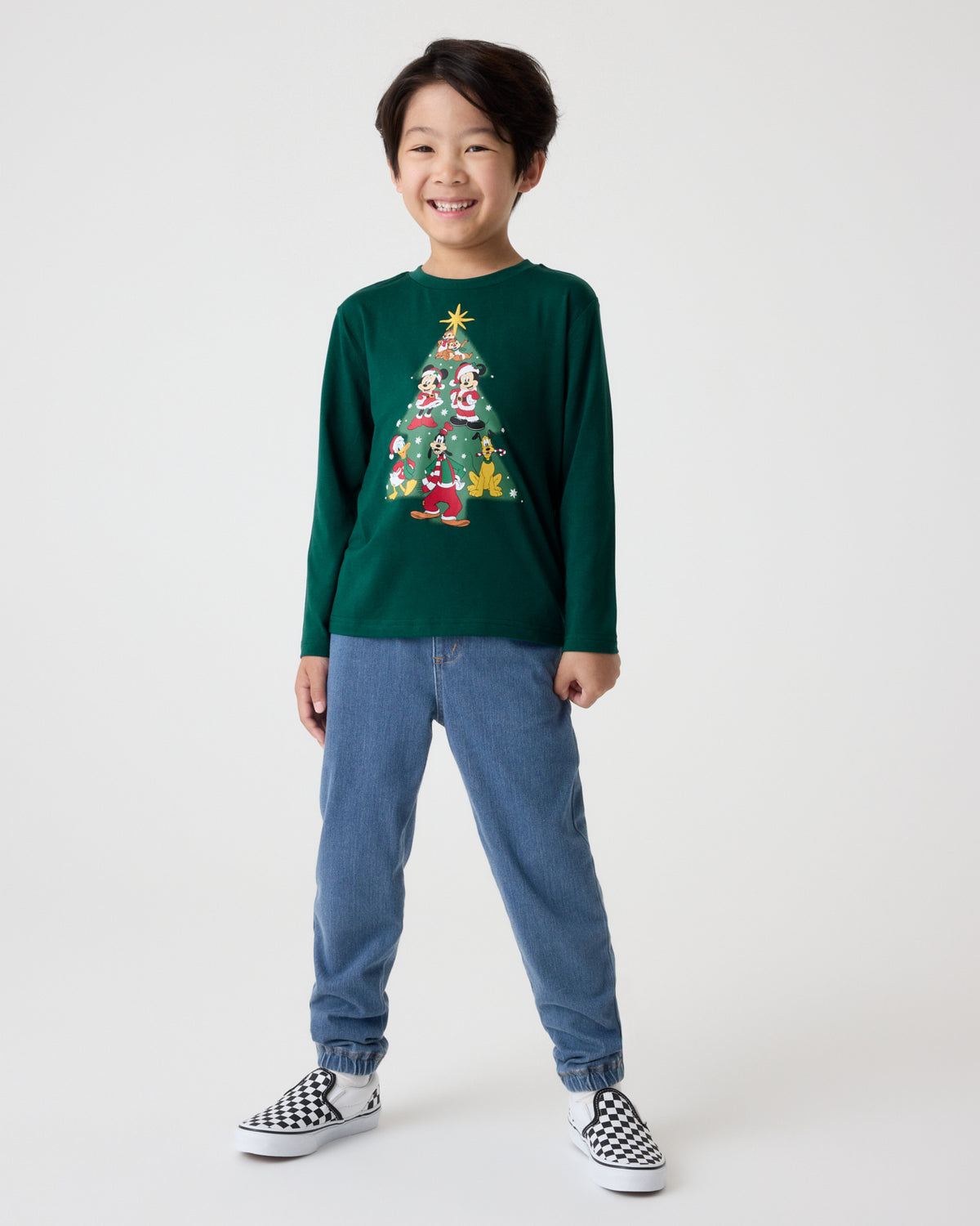 A smiling young boy stands against a light gray studio background wearing a dark green long-sleeve crewneck sweatshirt with a colorful Christmas tree graphic on the front, blue jeans, and black-and-white checkered slip-on sneakers.