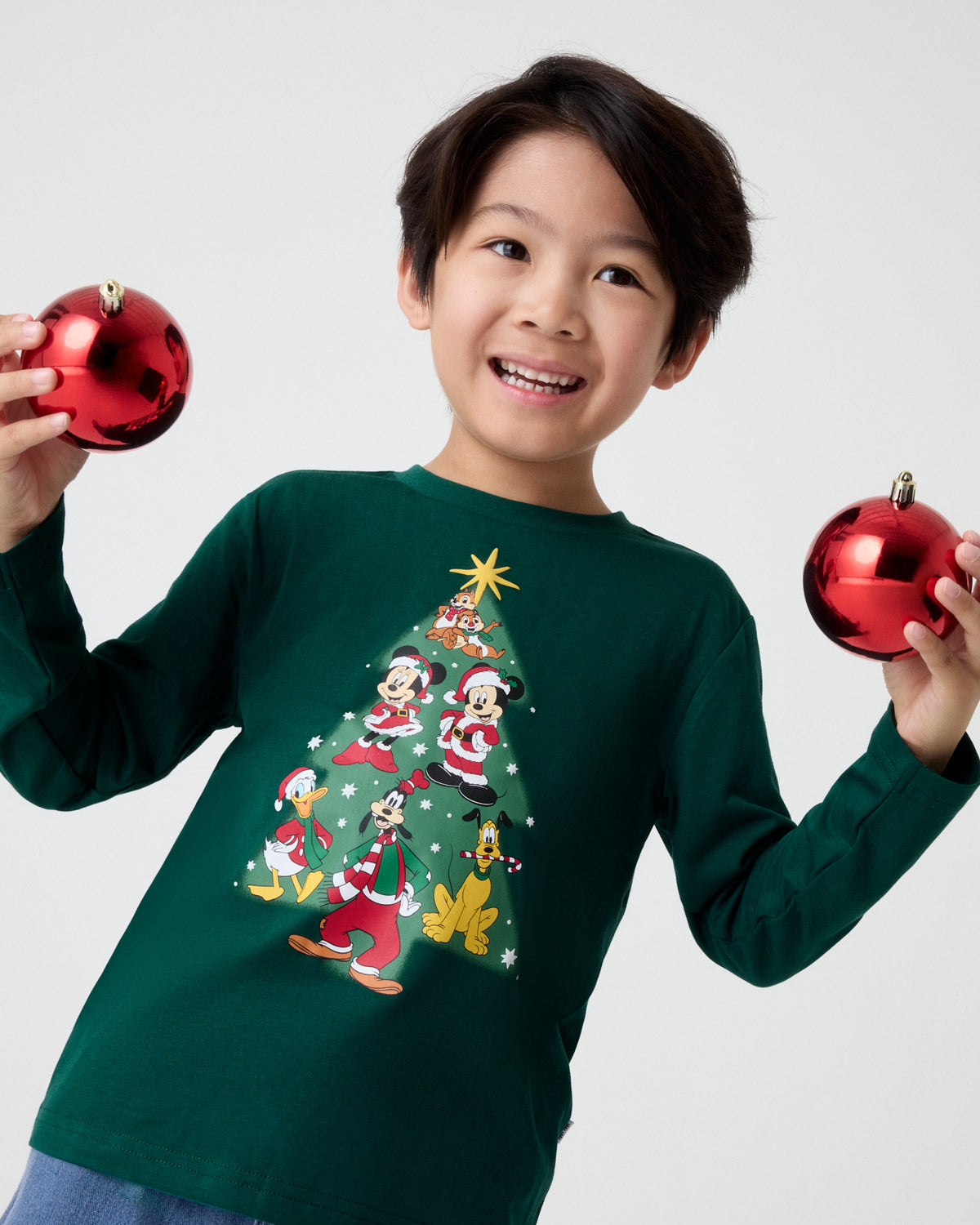 A smiling young boy wearing a dark green long-sleeve Christmas shirt with a Mickey Mouse and friends tree graphic, holding two shiny red ornaments against a light gray background.