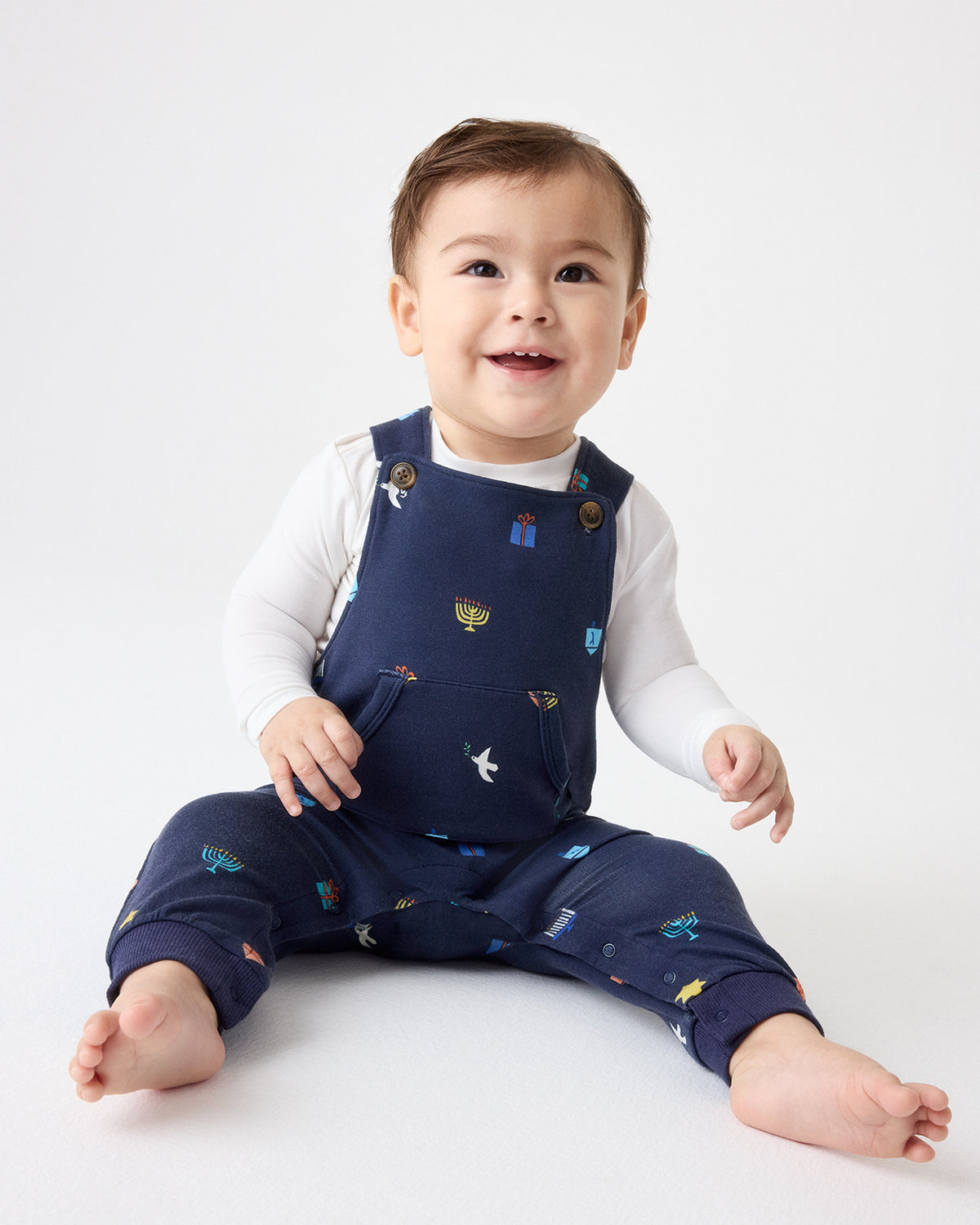 A smiling baby sitting on a white studio floor wearing navy blue overalls with colorful Hanukkah motifs, layered over a white long-sleeve bodysuit.