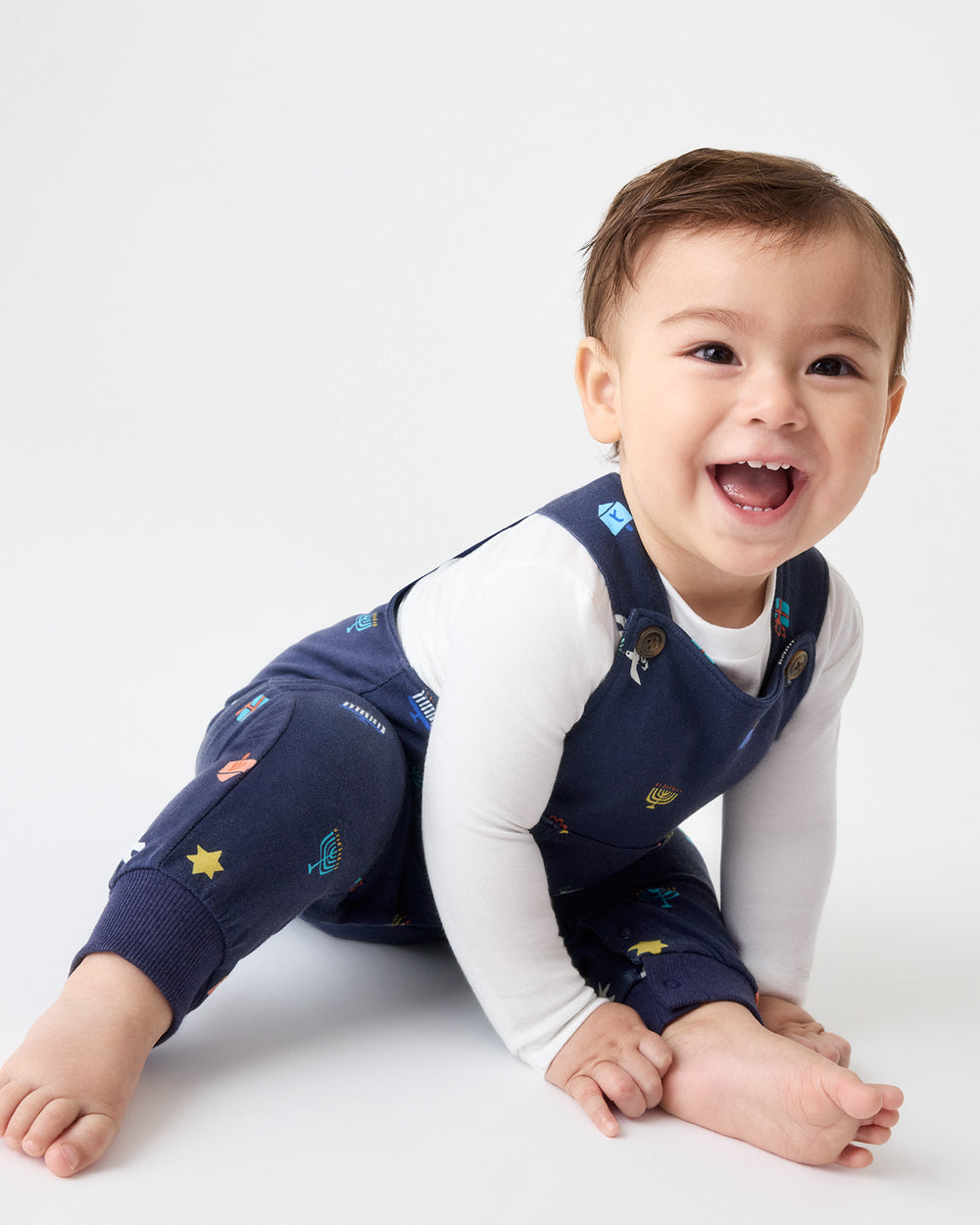 Crawling baby wearing navy blue overalls with colorful Hanukkah motifs, layered over a white long-sleeve bodysuit.