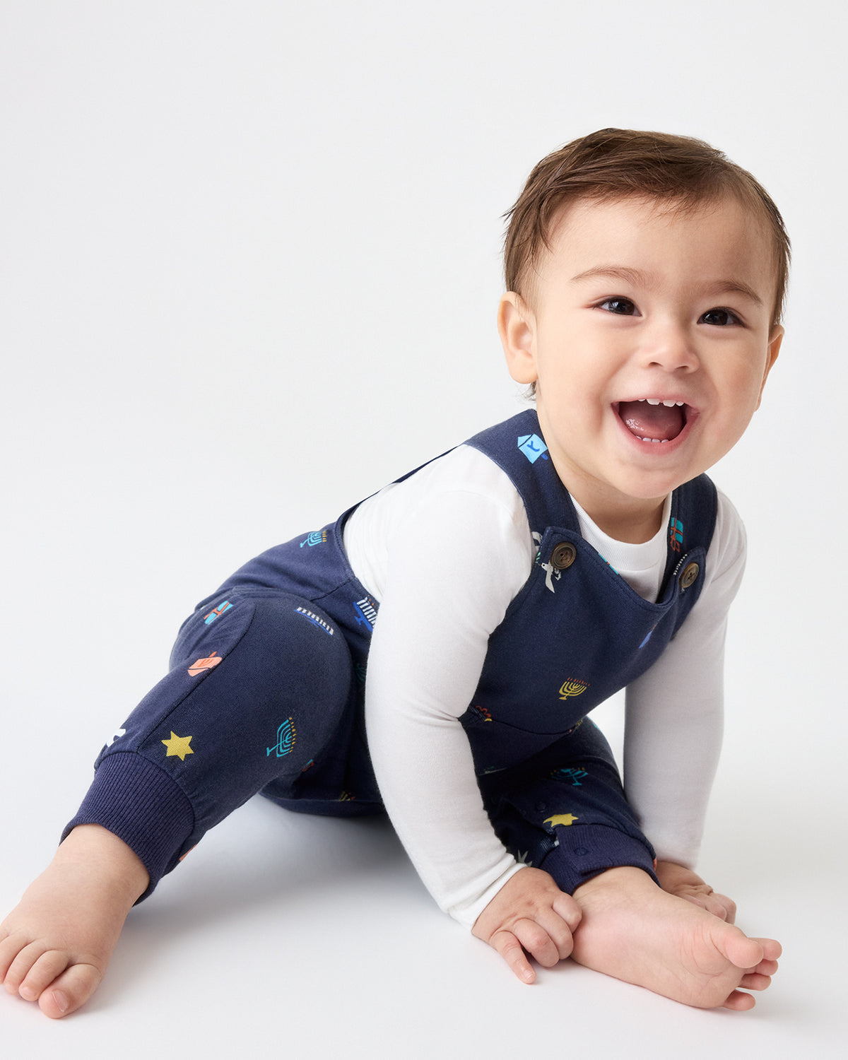 Crawling baby wearing navy blue overalls with colorful Hanukkah motifs, layered over a white long-sleeve bodysuit.