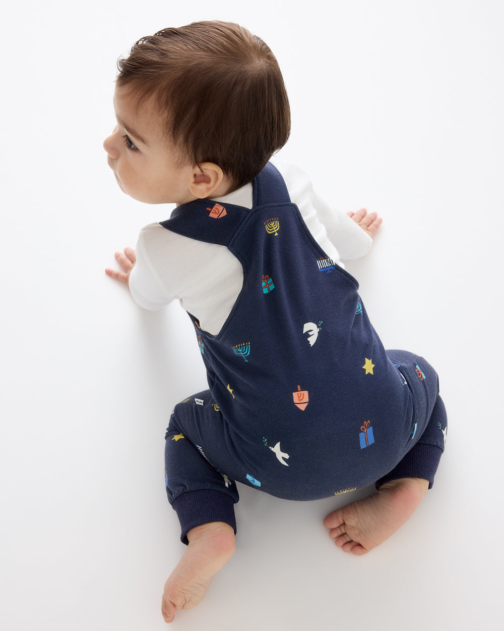 A baby wearing a navy blue romper with a colorful Hanukkah-themed print, sitting on a white studio background.