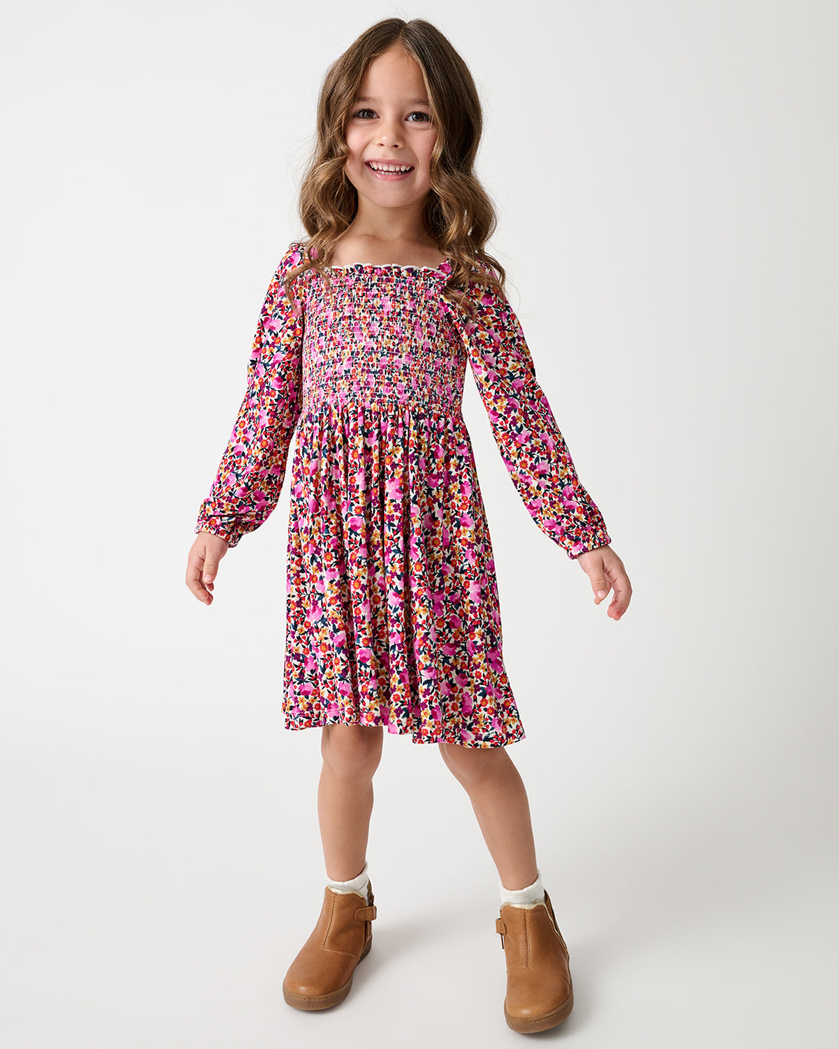 A young girl with wavy brown hair stands smiling in a pink, purple, and yellow floral smocked dress with long sleeves, paired with white socks and brown boots, against a light gray studio background.
