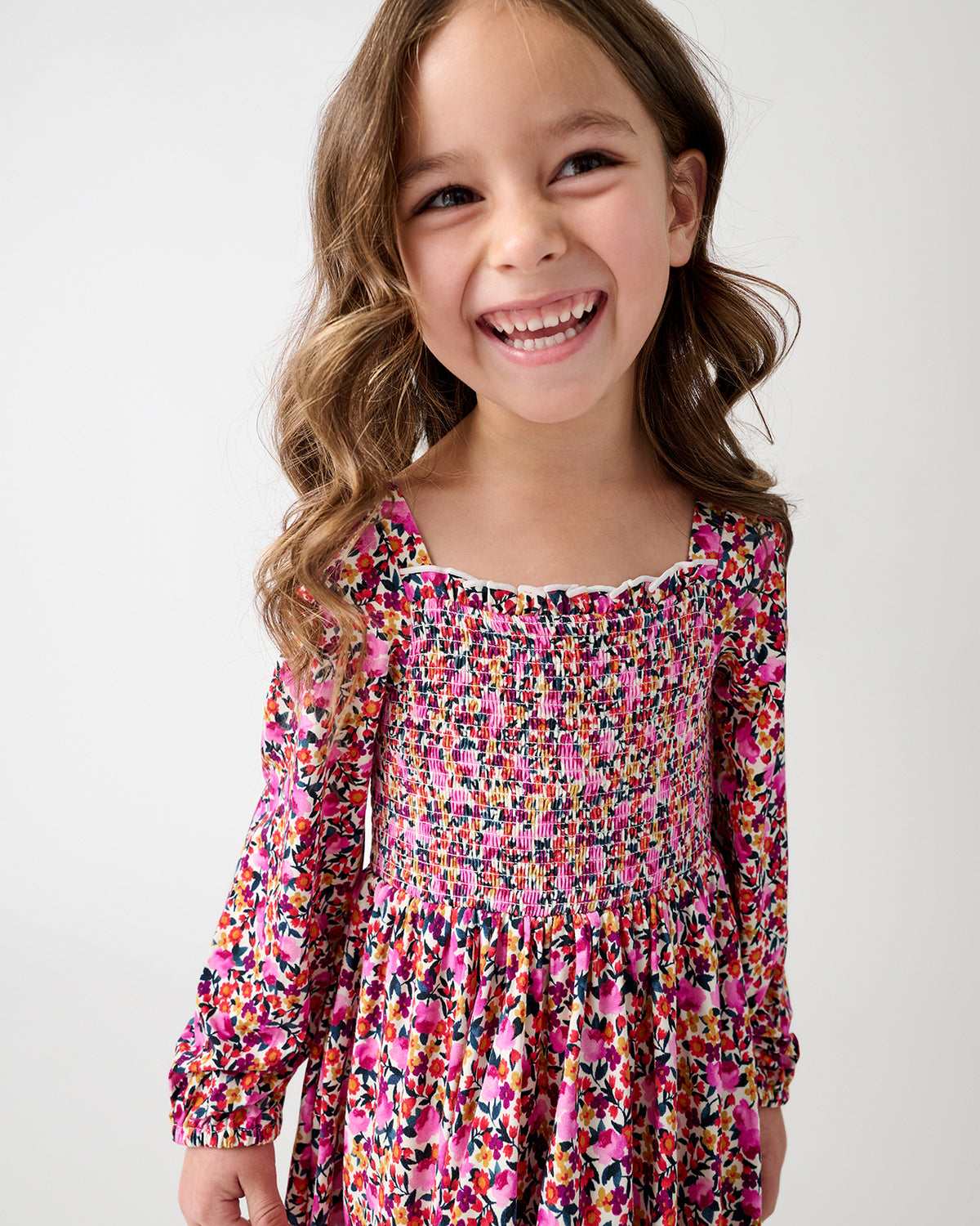 A young girl with long brown hair smiling, wearing a long-sleeve smocked dress with a vibrant multicolor floral pattern in pink, orange, yellow, and blue on a light background.