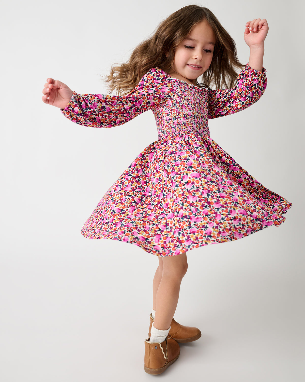 A young girl is twirling in a long-sleeved, smocked-bust dress with a colorful floral print. She is wearing brown boots and white socks against a plain light gray studio background.