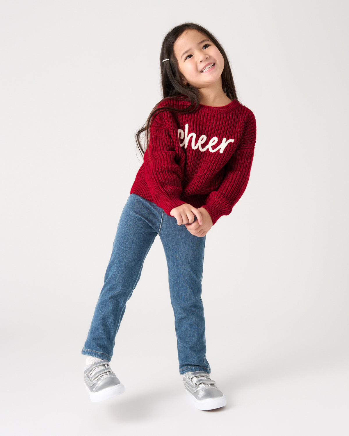 A young girl smiling and posing in a red ribbed knit sweater with 'cheer' written in white script across the chest, paired with blue jeans and silver sneakers, against a plain white studio background.