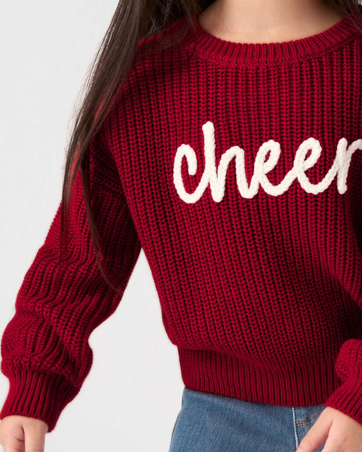 A close-up of a person wearing a chunky knit crewneck sweater in deep burgundy red with the word 'cheer' written across the chest in white script, paired with blue jeans.